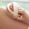 An exfoliating brush with soft massage nodules featuring a wooden base and a fabric strap, being held against skin.
