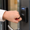 A hand wearing a brown ceramic ring is tapping a contactless access card reader on a door.