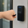 A hand wearing a black NFC carbon fibre ring interacts with a card reader mounted on a wall.