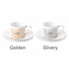 A ceramic coffee mug set featuring a golden and a silvery mug with matching saucers. Each mug has a decorative design.
