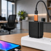 A black USB data blocker with an orange connector plugged into a power adapter, next to a smartphone on a desk.