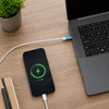 A smartphone charging beside a laptop, connected by a white USB cable on a wooden desk, with a green charging indicator.