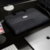 A dark grey laptop bag rests on a desk beside a computer and phone, featuring a handle and a logo.