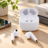 White half in-ear design earbuds rest beside their charging case on a wooden surface, with a smartphone and plant in the background.