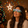 Paper diffraction glasses in blue with festive designs, worn by a woman against a background of Christmas lights and decorations.