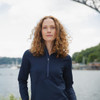 A woman stands by the water wearing a navy half-zip top. The image features a scenic background with a bridge.