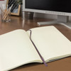 A large, soft cover Moleskine notebook in cream with lined pages and a purple ribbon bookmark, placed on a desk.