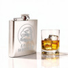 A stainless steel hip flask sits beside a glass of whiskey with ice, featuring an engraved design and a logo.