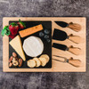 A bamboo cheese board featuring assorted cheeses, fruits, nuts, and four wooden handled cheese knives.