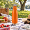 A drink bottle in vibrant orange, made of stainless steel, sitting on a picnic blanket with food and drinks nearby.