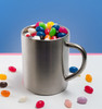 A silver mug filled with assorted colourful jelly beans, surrounded by a few scattered beans on a white surface.