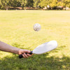 A pickleball paddle in hand and a perforated pickleball hovering above a grassy field.