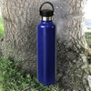 A drink bottle in vibrant blue stainless steel with a black lid and handle, set against a tree trunk.
