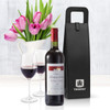 A black wine carrier beside a bottle of red wine and two glasses filled with wine, with tulips in the background.