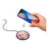 A wireless charging pad in black with a pink and grey design, alongside a smartphone being charged.