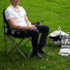 An oversized folding chair in grey, featuring a man seated with a drink. Nearby, a cooler bag and glasses are visible.