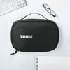 A black toiletry bag with a handle and a logo on the front, placed on a light wooden surface with office supplies.