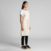 A linen apron in a light beige colour, worn over a white t-shirt and paired with black pants and shoes.