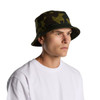 A camouflage bucket hat in green tones, featuring a branding logo, worn by a person facing to the side.