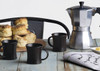 A silver espresso maker, three black mugs, and scones on a wooden table with a newspaper.