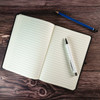 An A5 notebook with lined pages, featuring a black cover, a pen, and a blue pencil on a wooden surface.
