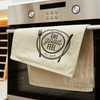 A cotton tea towel in cream with a black logo, hanging on an oven door.