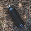 A drink bottle in matte black with a silver cap, featuring a logo. Set on a bed of pine needles.