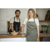 A green recycled apron worn by a smiling woman in a kitchen, with a man in a black apron in the background.