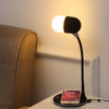 A black and white wireless charging sound lamp with a flexible neck, next to a smartphone on a charging pad.