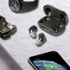 Wireless earbuds in grey resting on a marble surface next to a smartwatch and a smartphone, featuring a logo on the case.