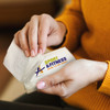 A hand holds a pack of tissues with a logo, featuring white tissues and a textured pattern. The pack has a colourful branding design.