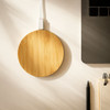 A round bamboo wireless charger in a natural finish, placed beside a laptop and stationery on a desk.