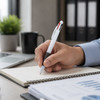 A white plastic pen with red and black accents rests near a notepad on a desk. A coffee mug and plant are in the background.