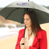 A grey umbrella with a logo, held by a woman wearing a red coat, set against a beach backdrop.