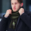 A man wearing a dark green ribbed scarf, layered under a black coat, amidst a snowy backdrop. The scarf features a logo.