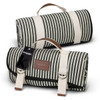 A striped picnic blanket in black and cream, folded with handles and a tag. It features a brown leather label.