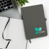 A grey notebook with a logo on the cover, lying open beside a laptop and a small plant.