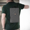 A grey laptop sling bag is worn on the back of a person against a white wall. It features a logo.