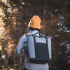 A grey and black cooler totepac is worn on the back of a person, surrounded by a forest backdrop.