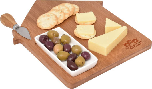 A wooden cheese board in the shape of a house with assorted cheeses, olives, crackers, and a knife.