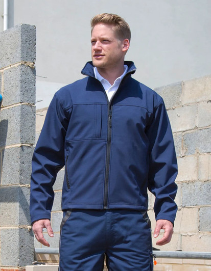 A man wearing a navy soft shell jacket stands against a partially constructed wall, with a branding logo visible.