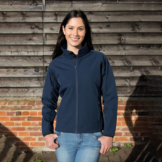 A navy softshell jacket with a zip, worn by a woman standing against a wooden wall. The jacket has a logo.