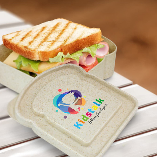 A sandwich box in a beige colour, featuring a logo and containing a sandwich with layers of ham, cheese, and lettuce.
