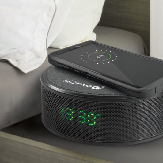 A wireless charger with a dark grey speaker design, featuring a phone on top and a digital clock display.