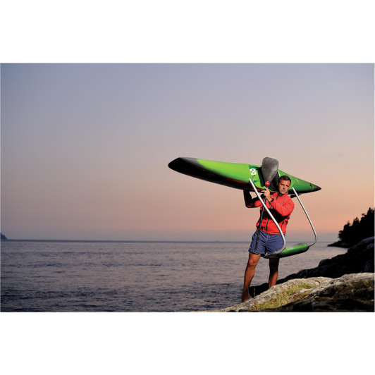 A man in a red top and shorts carries a green kayak over his shoulder by the water at sunset.