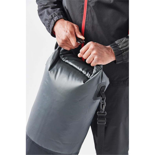 A close-up of a person holding the top of a grey roll-top backpack with a strap, featuring a logo.