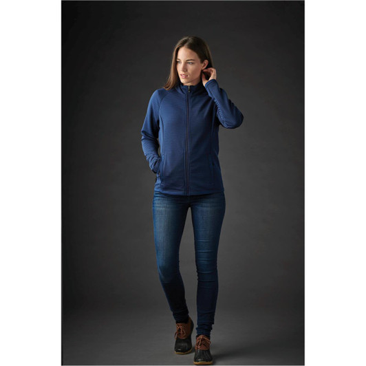 A woman models a navy blue jacket with a zip front and fitted design, paired with denim jeans. The background is dark.