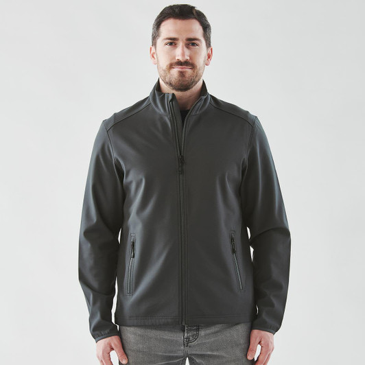 A man wearing a dark grey softshell jacket with a zip front and side pockets. The jacket has a logo.