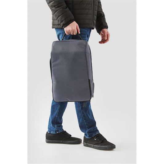 A person carrying a grey Toluca Backpack with a handle, wearing dark jeans and black shoes. The backpack has a logo.