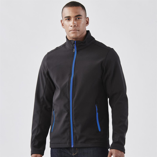 A man wearing a black softshell jacket with blue accents and zippered pockets, featuring a logo.